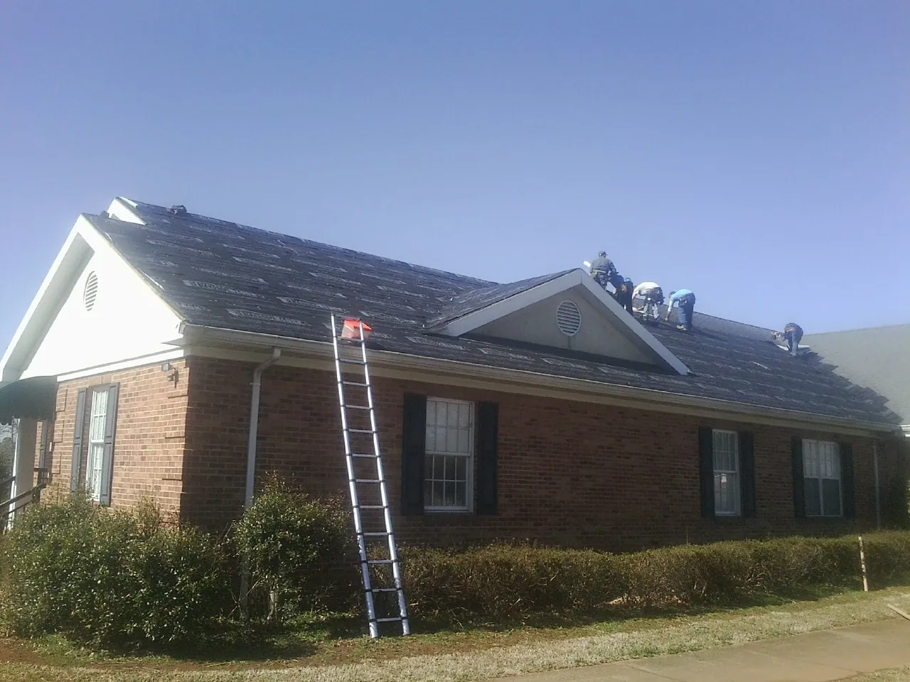 Crew tearing shingles off a brick ranch roof with a ladder set — Emergency Roof Repair work by our crew in Rocky Mount