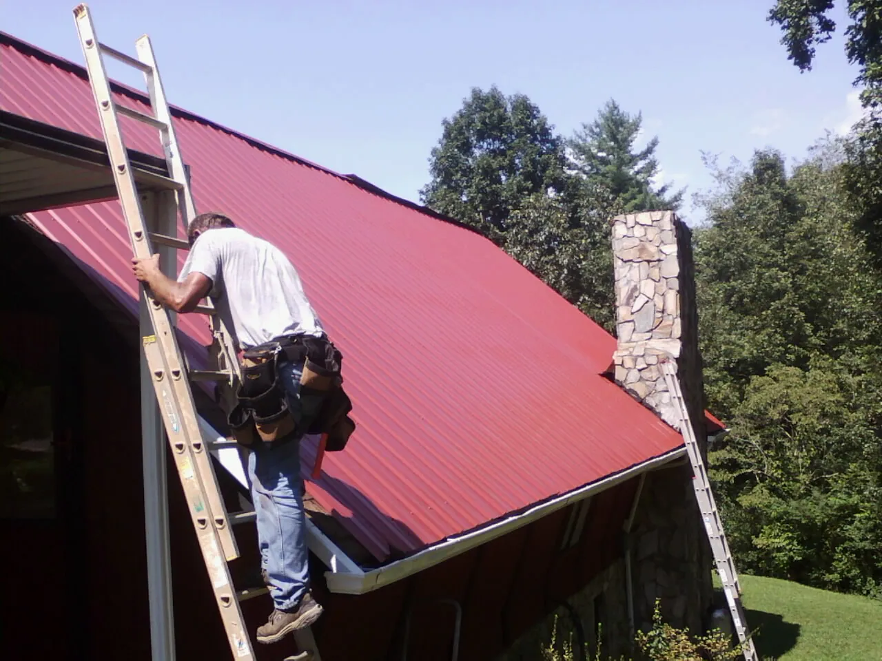 Roofer on a ladder beside a red metal roof and stone chimney in Rocky Mount
